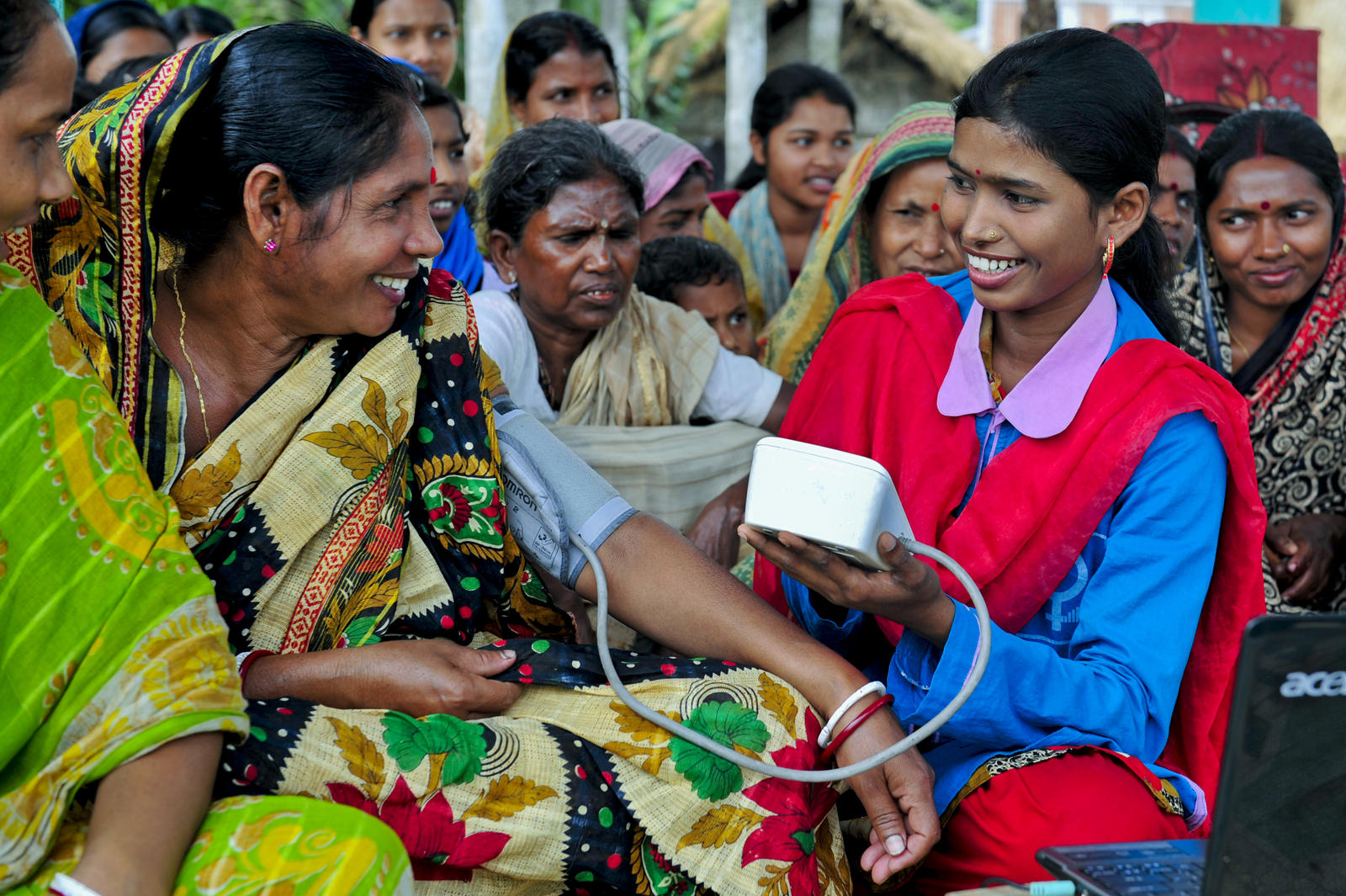 A woman in medical uniform is checking the blood pressure of other woman and there are other women sitting in the background