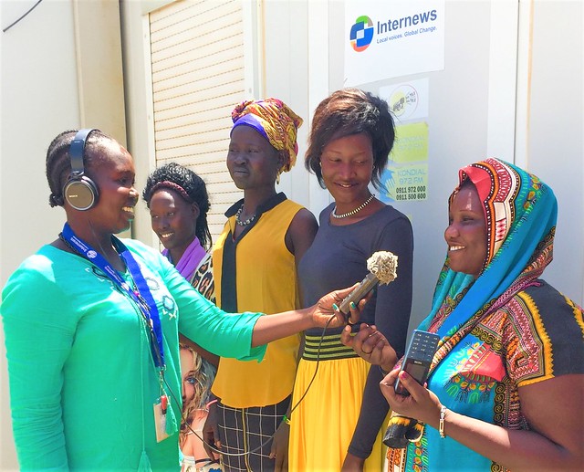 A young woman media person (wearing headphone and holding a audio recorder) is taking interview of other young woman. There are 3 other young girls standing behind them. All of them are smiling. Internews; March 2019; Bentiu, South Sudan