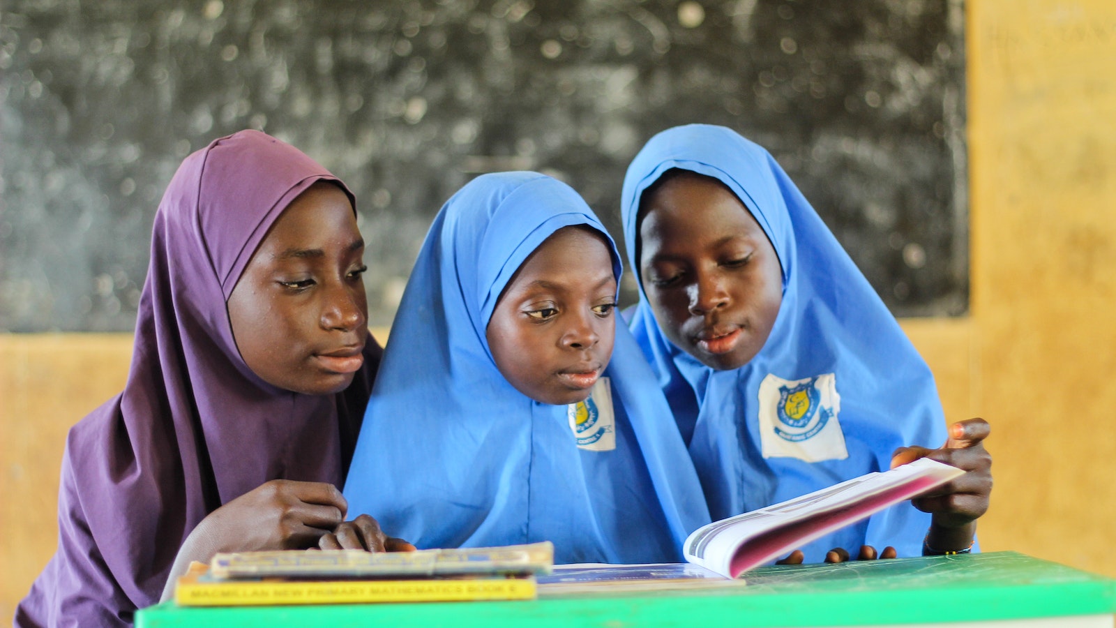 A Pair of Girls Wearing Blue Hijabs Reading a Book Beside a Young Woman Wearing Purple Hijab