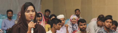 Young woman wearing a head covering and speaking into a microphone with other young people seated in the background