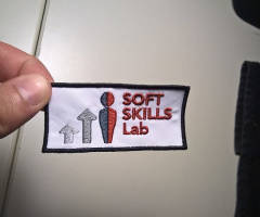 A hand holding a white patch with black outline that says SOFT SKILLS Lab in red embroider